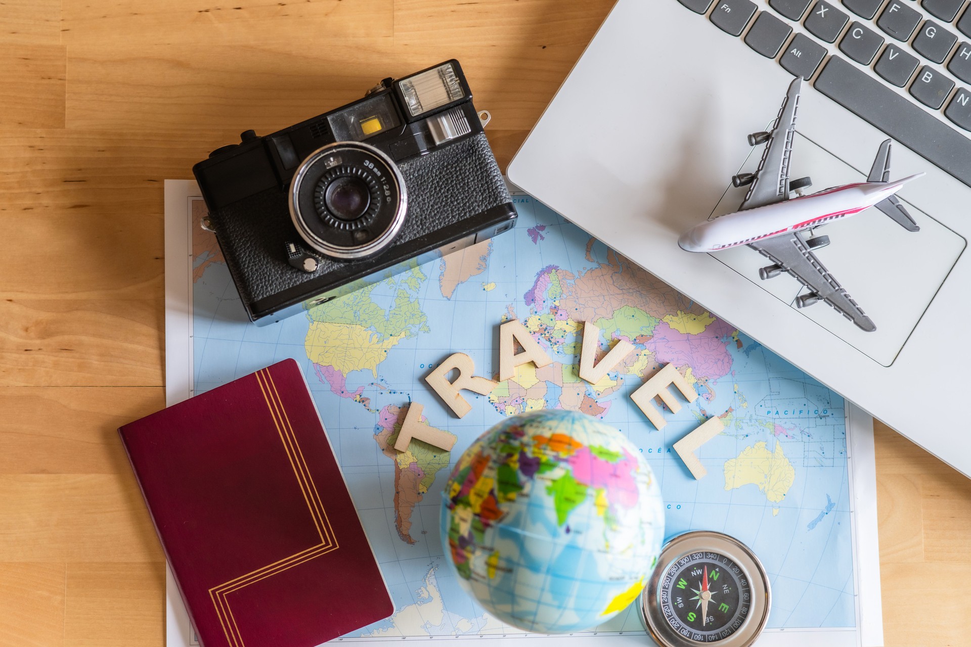 World map with globe, compass, passport, camera and laptop displaying a miniature airplane on top of a wooden table suggest planning and booking the next summer vacation trip